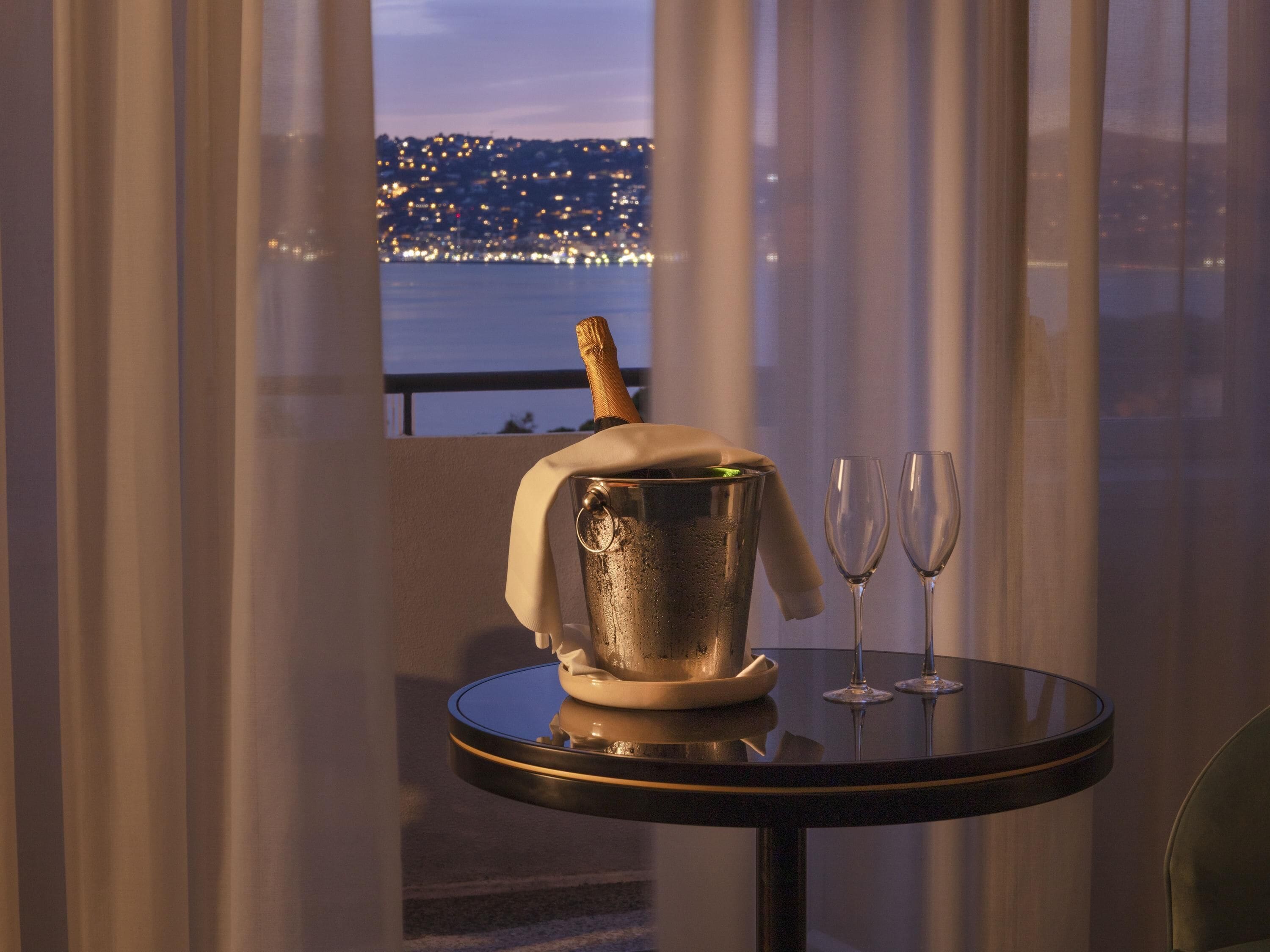 Ein Tisch mit einem Champagnerkühler und zwei Gläsern, einer Champagnerflasche und Blick auf eine Stadtlandschaft im Abendrot.