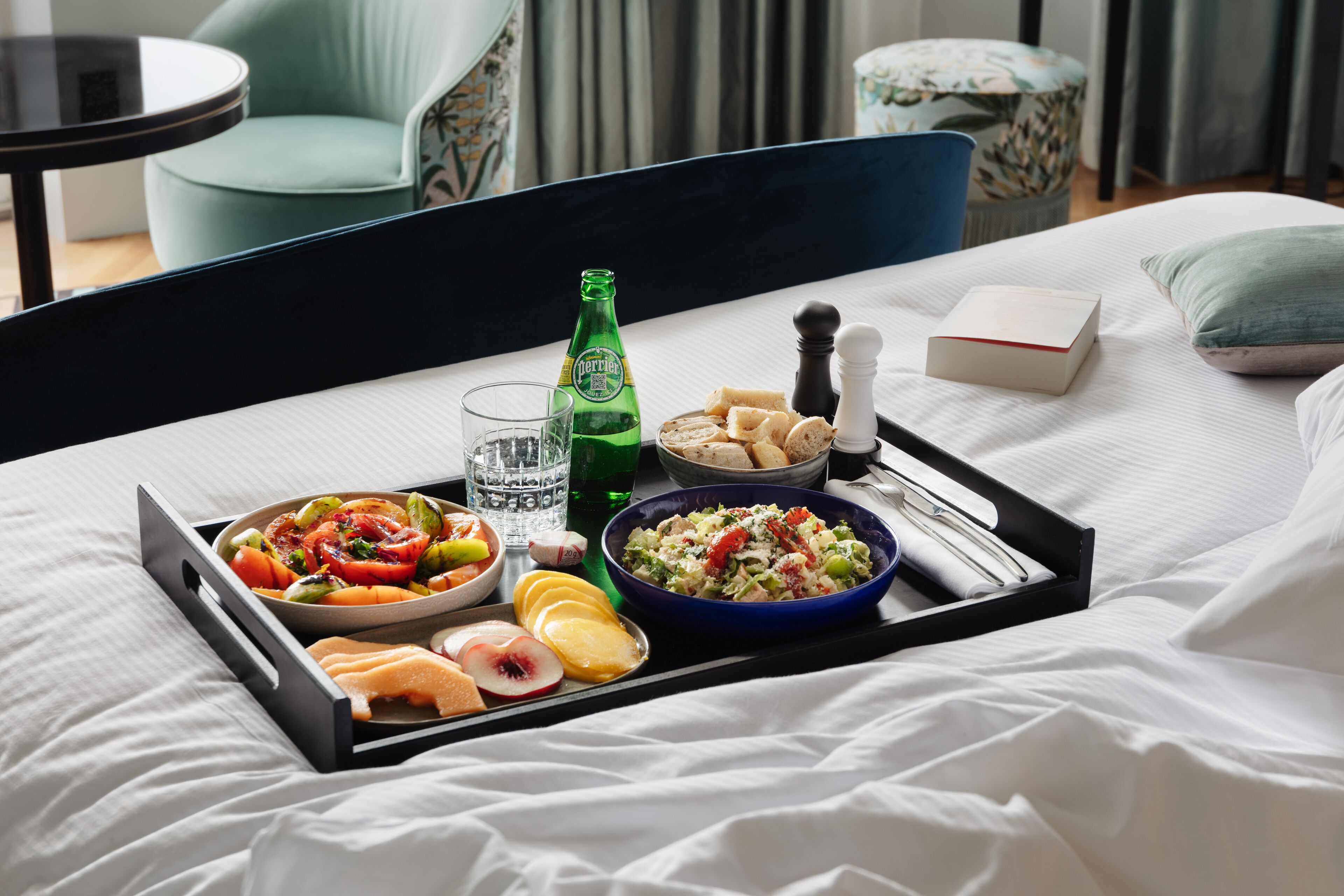 Ein ordentlich angerichtetes Frühstücksbrett auf einem Bett mit verschiedenen Früchten, einer Salatschüssel, Brot und einer Bierflasche.