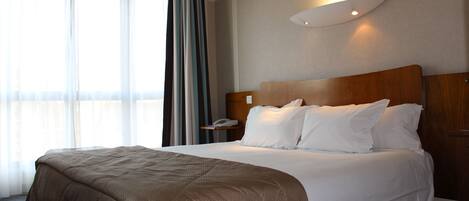 Habitación de hotel con una cama grande, una mesita de noche con lámpara y vistas al exterior a través de cortinas translúcidas.