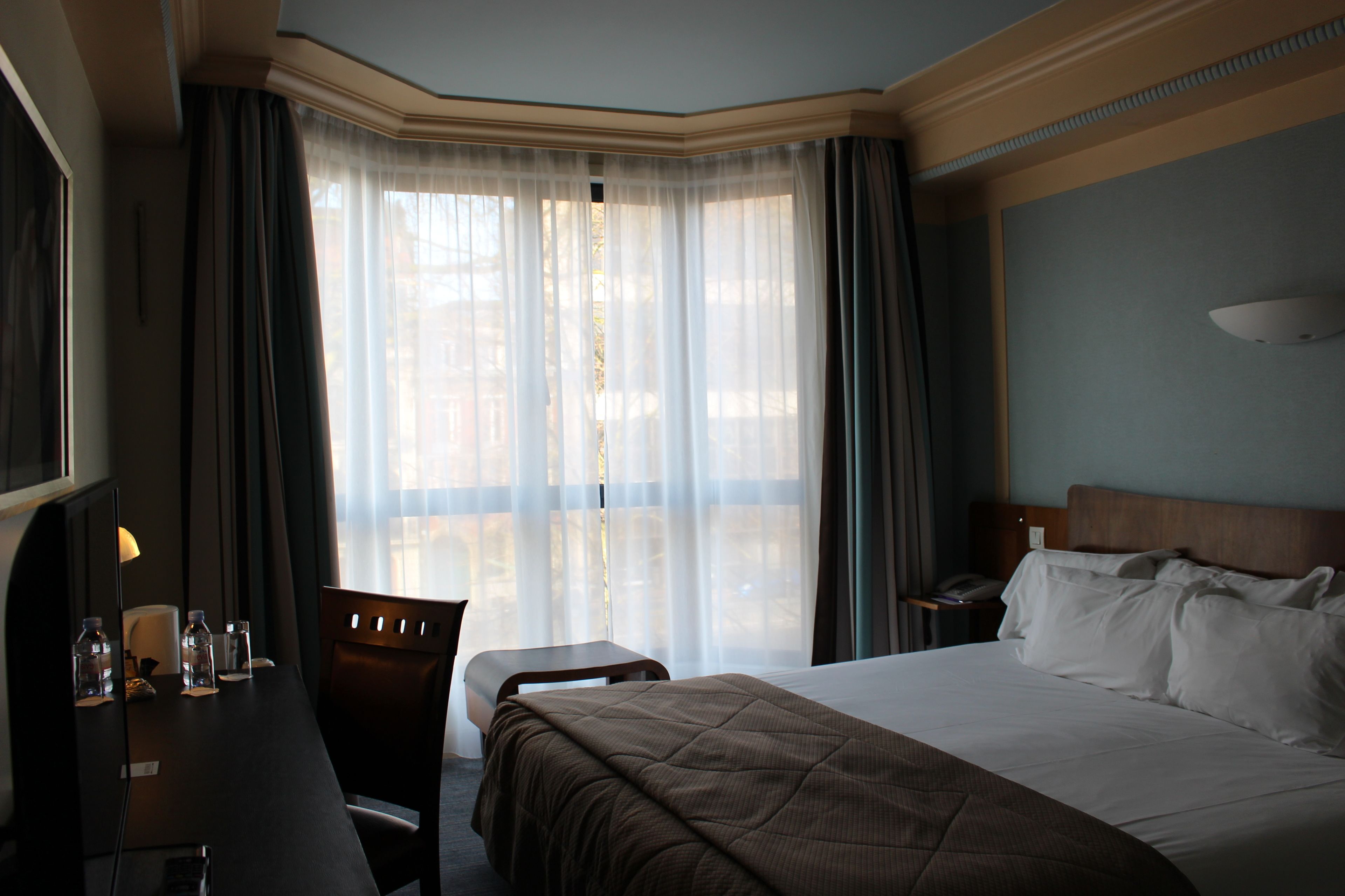A hotel room with a large bed, a desk with a chair, a lamp, and a window with sheer curtains.