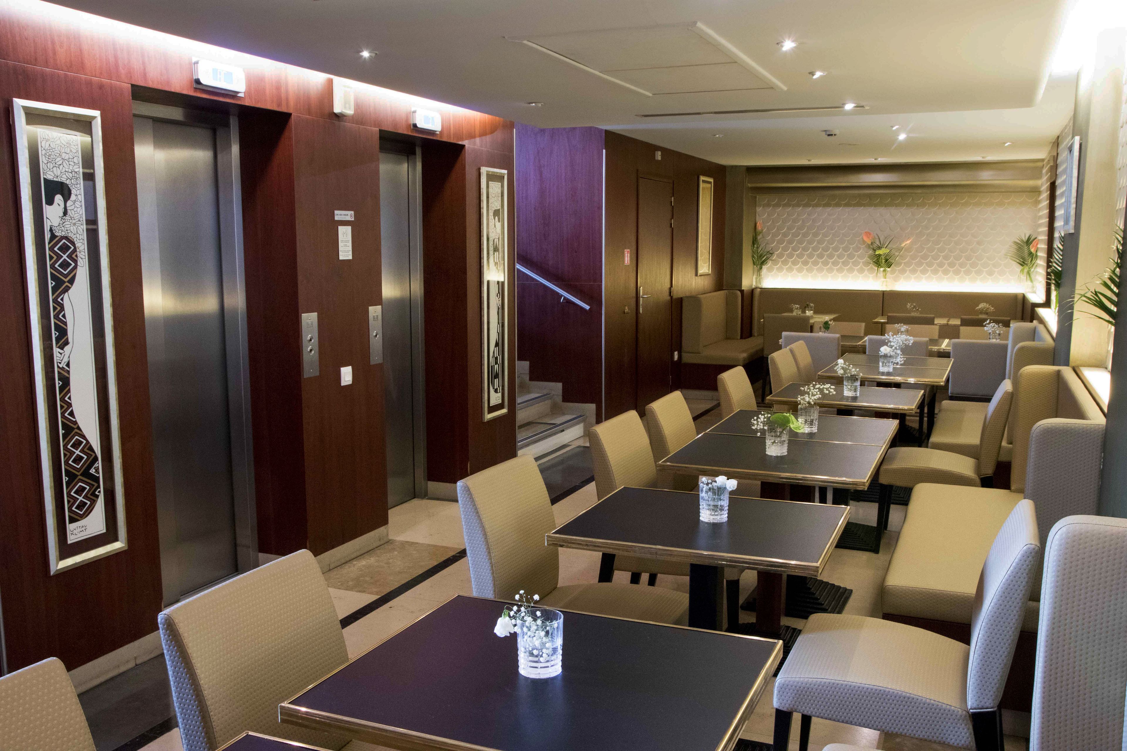 Een restaurantgedeelte van een hotel met meerdere tafels en stoelen, een lift en een muur met een geometrisch patroon.