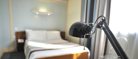 Quarto de hotel com cama, luminária de mesa e janela com cortinas.