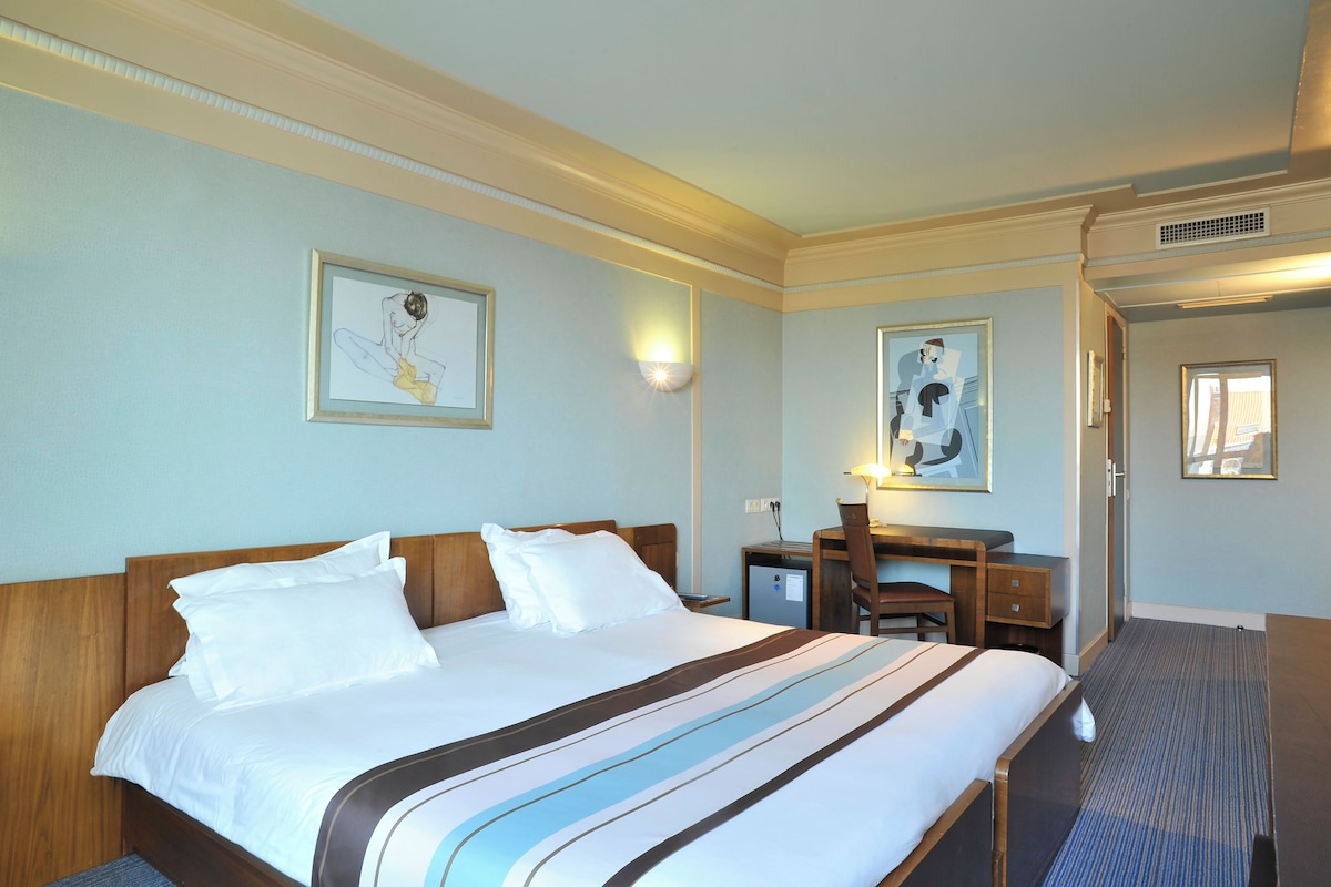 A hotel room with a large bed, a desk, and a chair. There are two framed pictures on the wall.