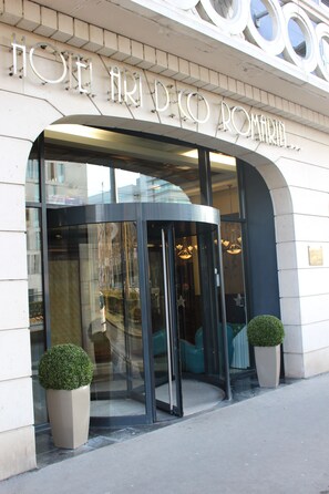 Entrada de hotel con puerta giratoria, fachada de vidrio y dos plantas en macetas.