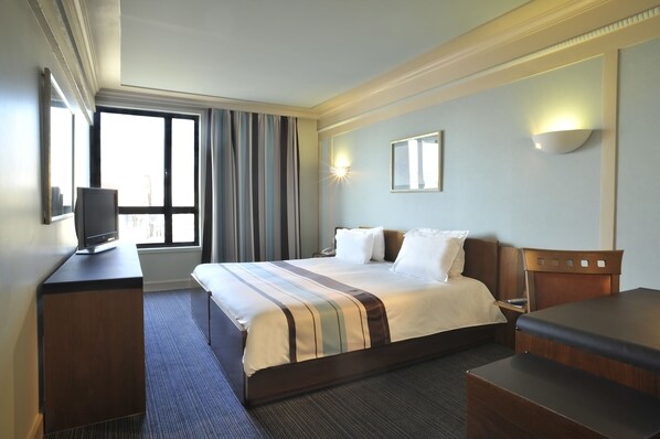 Una habitación de hotel con una cama grande, un escritorio, un televisor y una ventana con cortinas.