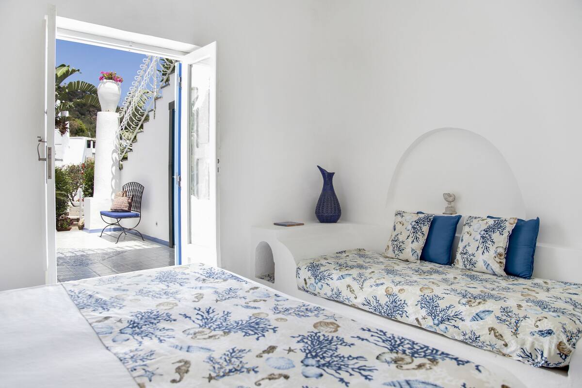 Ein Schlafzimmer mit einem Bett, einem Stuhl, einer Vase und Blick ins Freie.