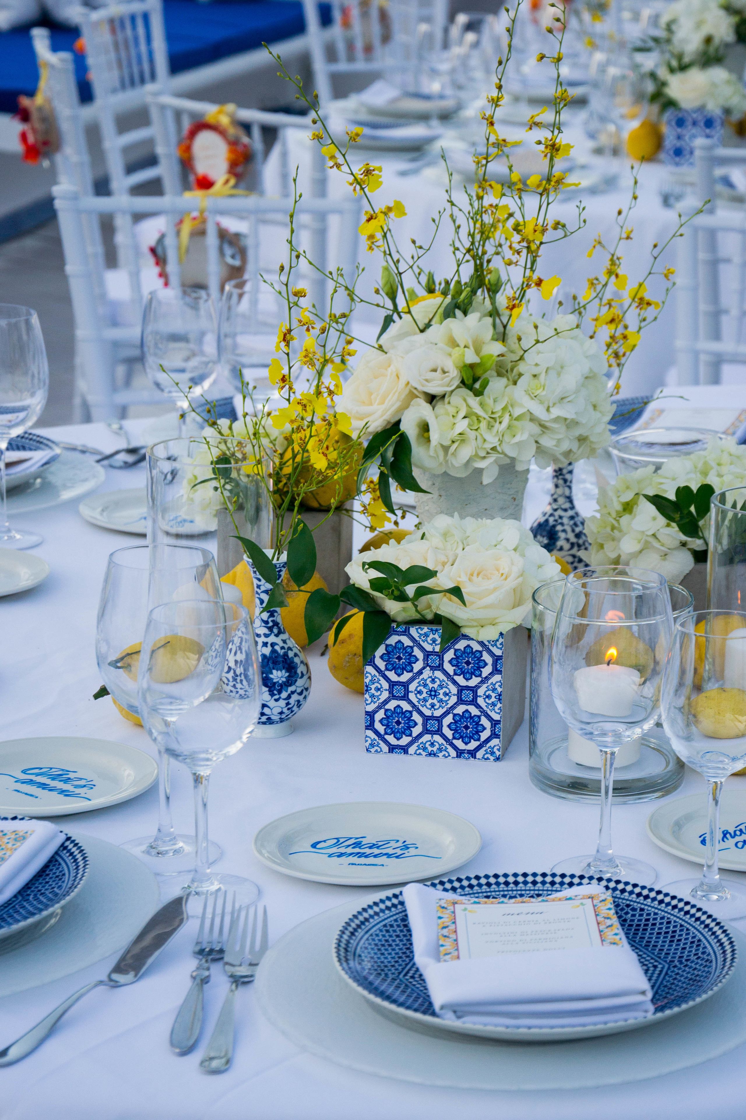 Una mesa dispuesta para un evento formal, con vajilla blanca y azul, cristalería transparente y centros de mesa florales.