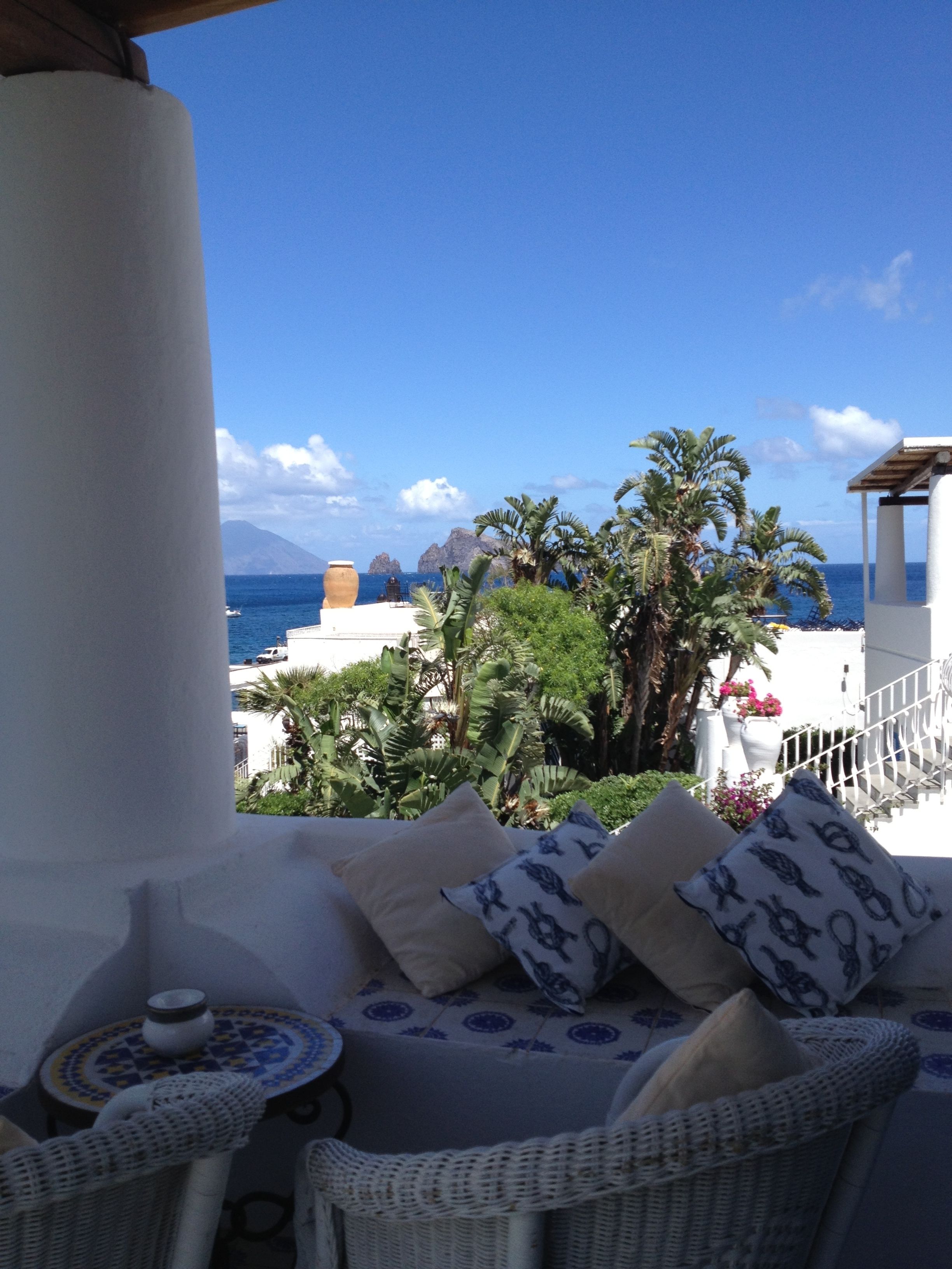 A balcony with wicker furniture, a view of the sea, and tropical plants.