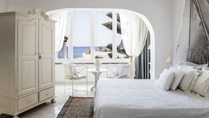 Une chambre avec un grand lit, une armoire blanche, une petite table avec des chaises et une vue sur la mer par une fenêtre en arc de cercle.