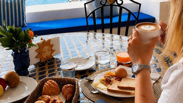 Une personne assise à une table, avec vue sur la mer, un livre et des éléments de petit-déjeuner.