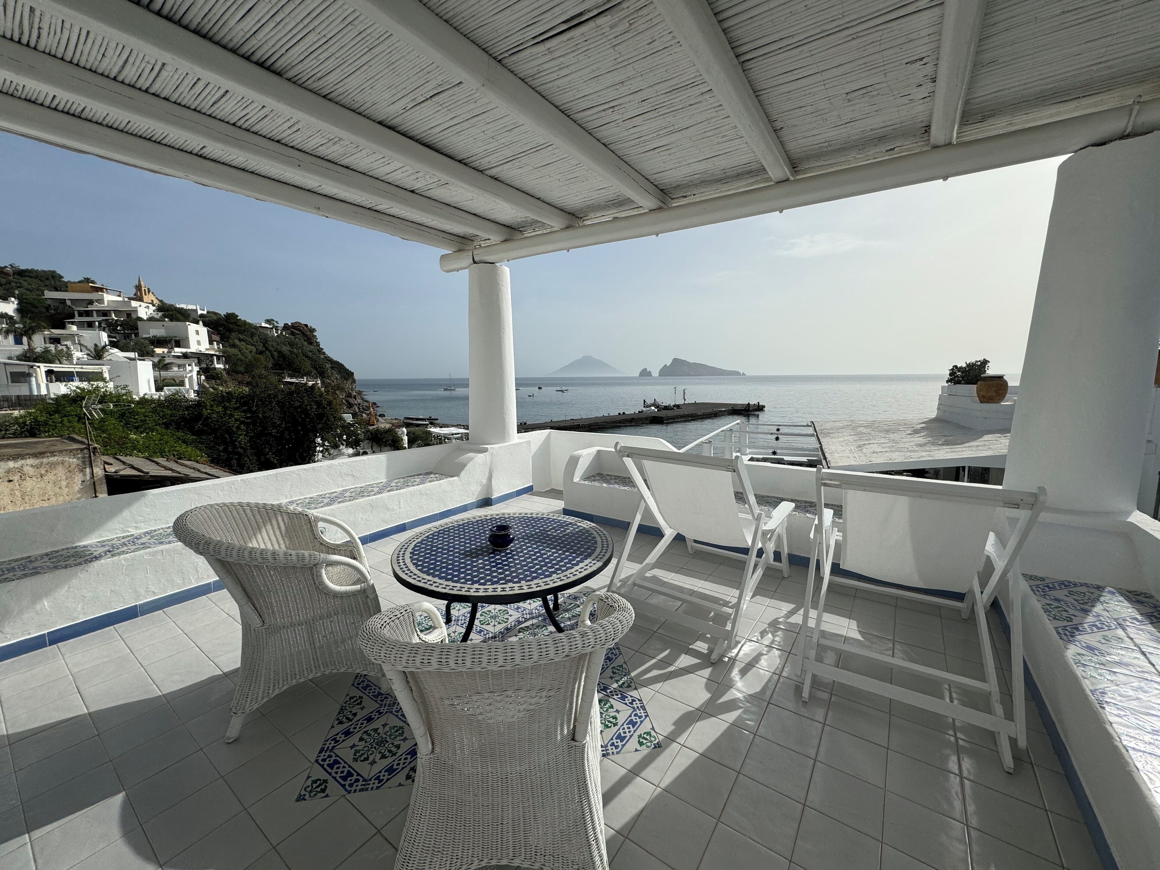 Una terraza con mobiliario de mimbre blanco, una mesa con diseño y vistas al mar y a la costa lejana.