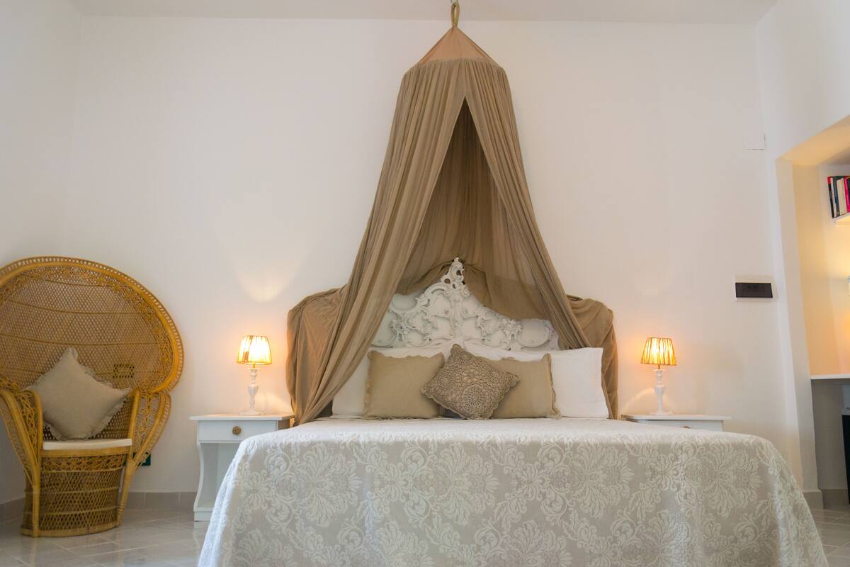 Ein Schlafzimmer mit Himmelsbett, Korbsessel, zwei Nachttischen mit Lampen und einem Bücherregal.