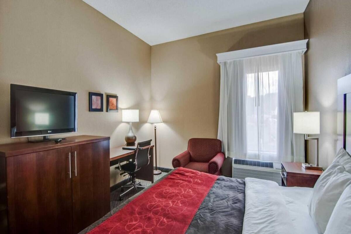 Een hotelkamer met een bed, een televisie, een bureau met stoel, een rode fauteuil en een raam met gordijnen.