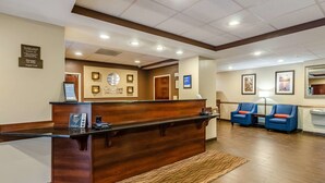 A hotel reception area with a wooden reception desk, a digital information board, and a seating area with blue chairs.