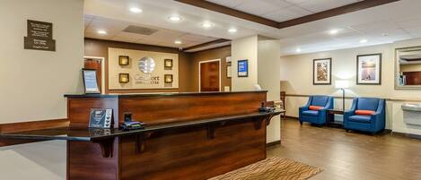 Zona reception di un hotel con una scrivania in legno, una bacheca digitale e un'area salotto con poltrone blu.