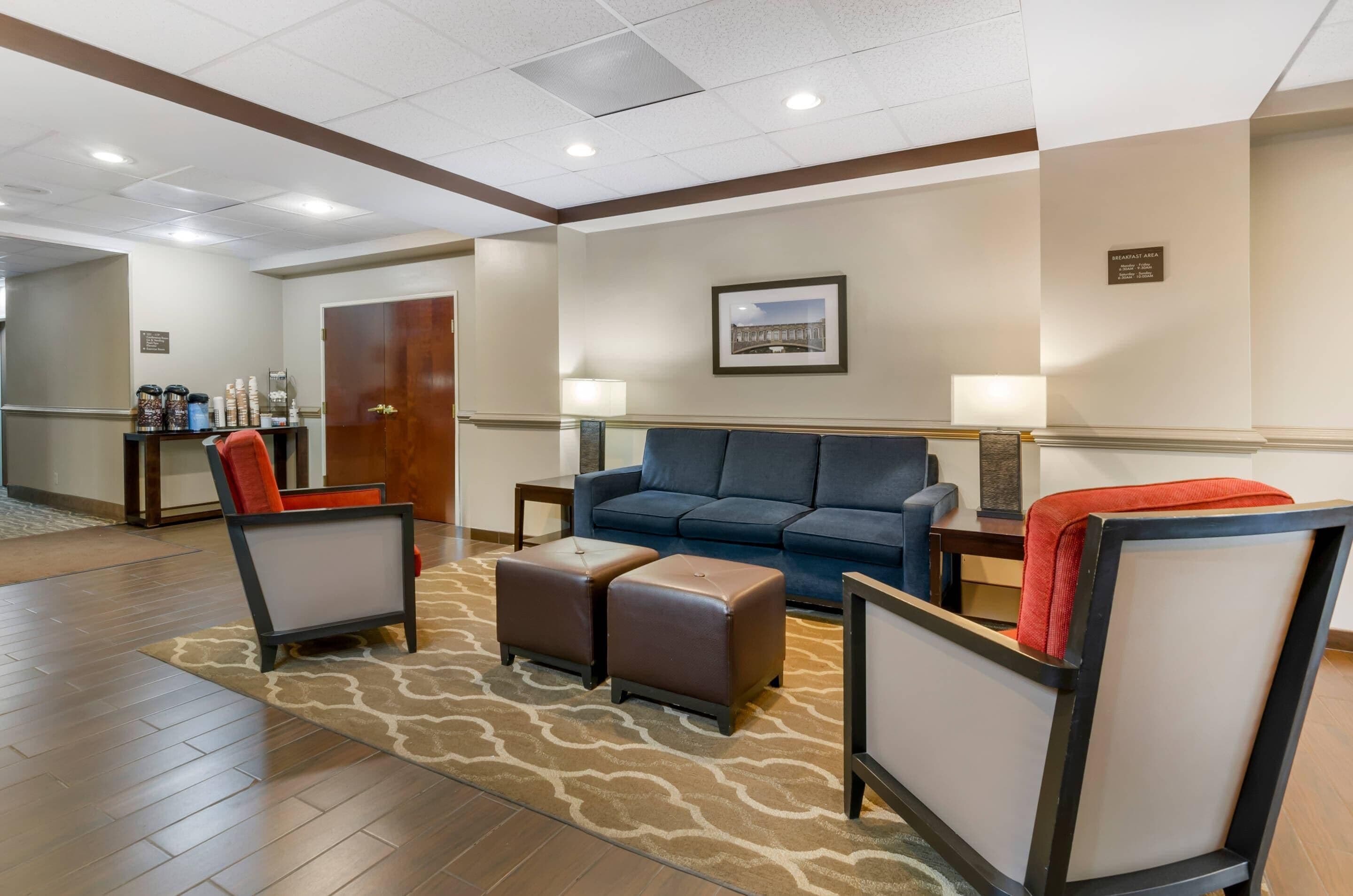 A hotel lobby with a blue sofa, two armchairs, a coffee station, and a framed picture on the wall.