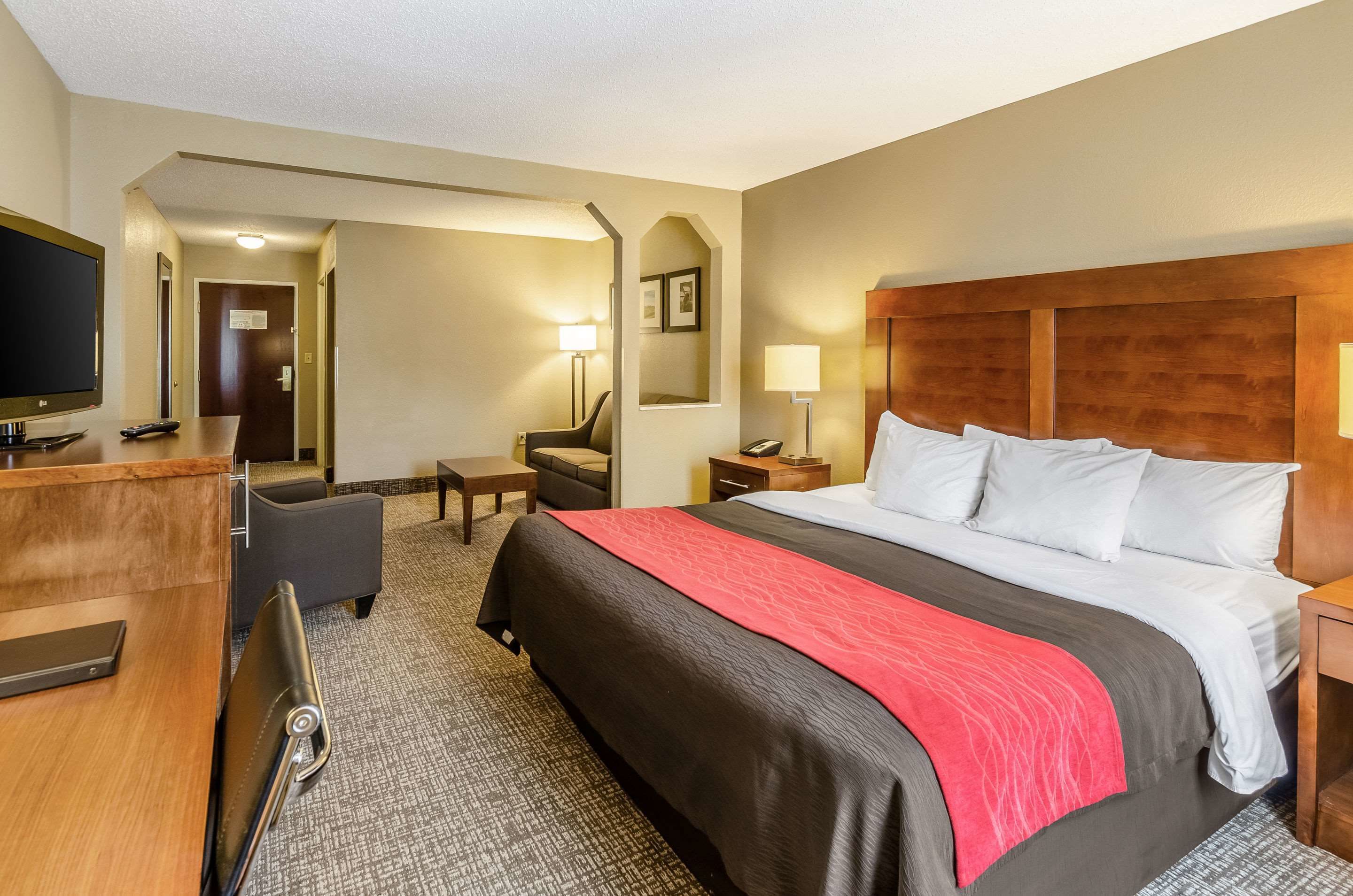 A hotel room with a large bed, a desk with a flat-screen TV, a chair, a desk lamp, and a bedside table.