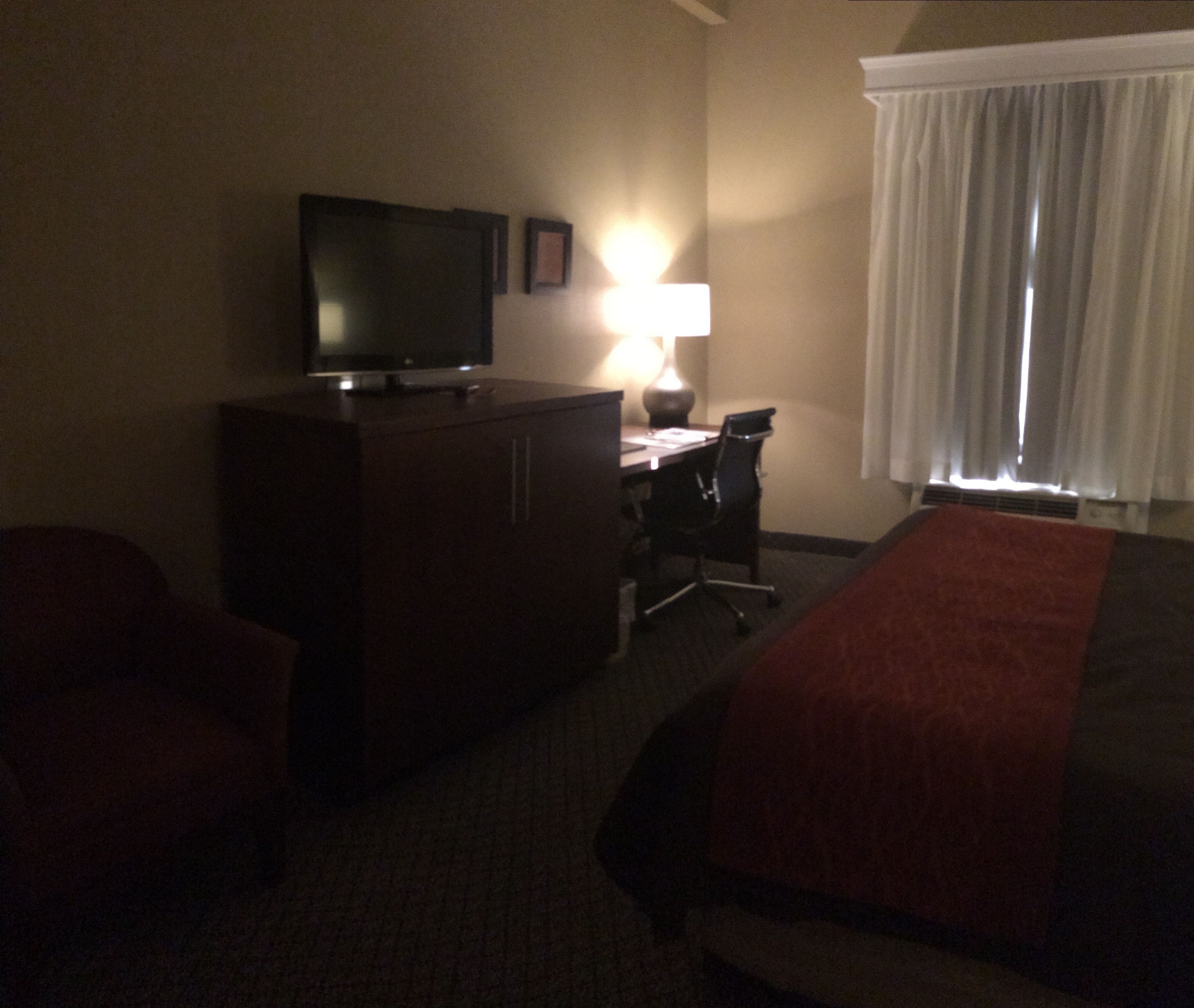 Quarto de hotel com cama, escrivaninha, cadeira, televisão e abajur.