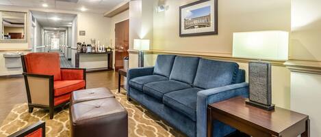 A hotel lobby with a blue sofa, red armchair, and a brown ottoman. A wooden table with a lamp is also visible.