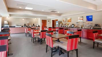Een restaurantgedeelte in een hotel met meerdere tafels en stoelen, een balie met een koffieapparaat en een koelkast.