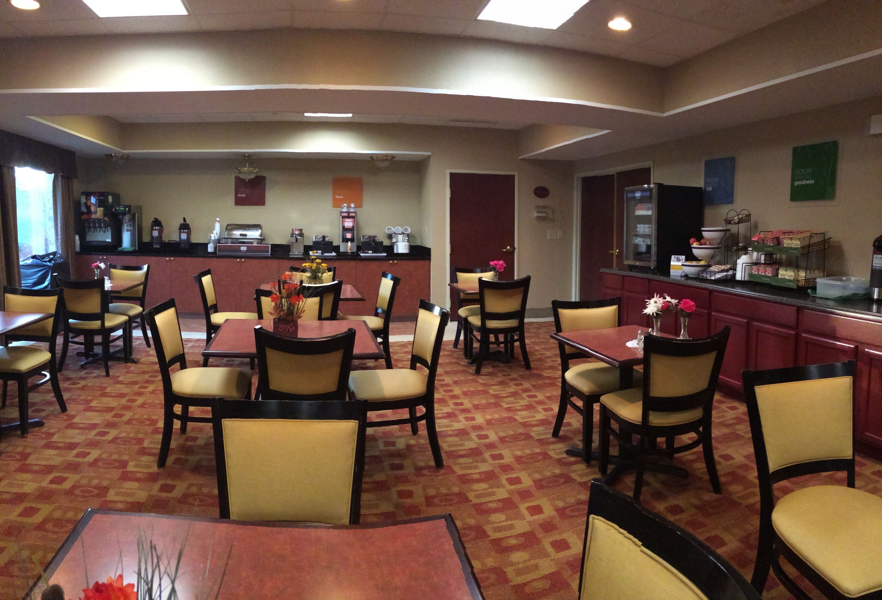 A hotel dining area with multiple tables and chairs, a counter with coffee machines, and a view of the kitchen in the background.