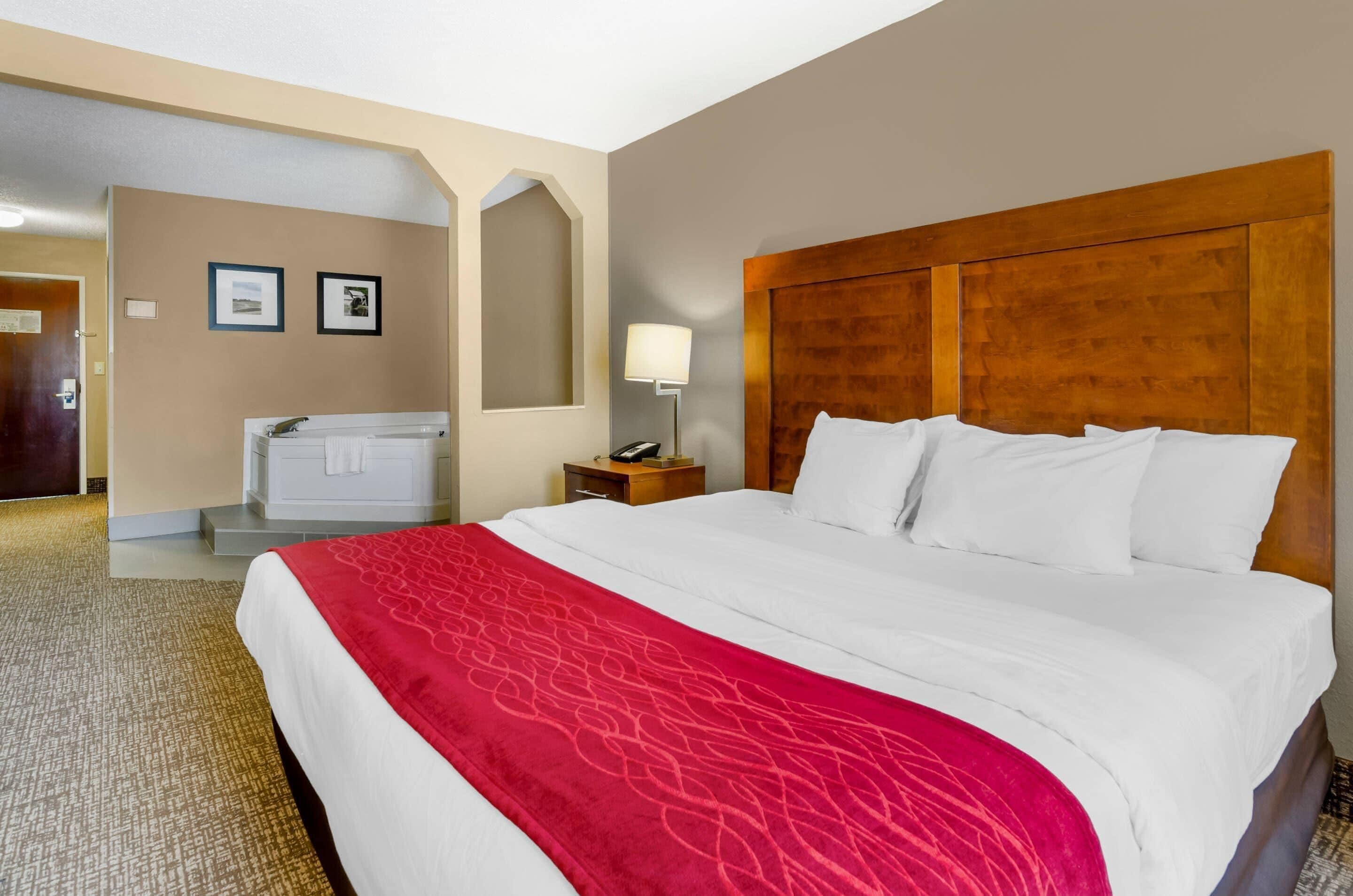 Habitación de hotel con una cama grande, cabecera de madera, manta roja, mesita de noche con lámpara y baño con bañera y toallero.