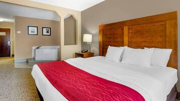 A hotel room with a large bed, a wooden headboard, a red throw blanket, a nightstand with a lamp, and a bathroom with a bathtub and towel rack.
