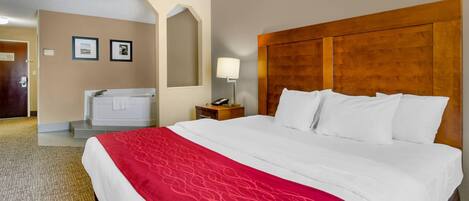 Habitación de hotel con una cama grande, cabecera de madera, manta roja, mesita de noche con lámpara y baño con bañera y toallero.