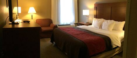 A hotel room with a bed, a desk with a computer and lamp, a chair, and a window with curtains.