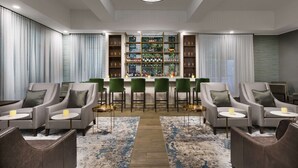 Un espace lounge moderne avec des fauteuils gris, un bar doté d’étagères remplies de bouteilles et un lustre.