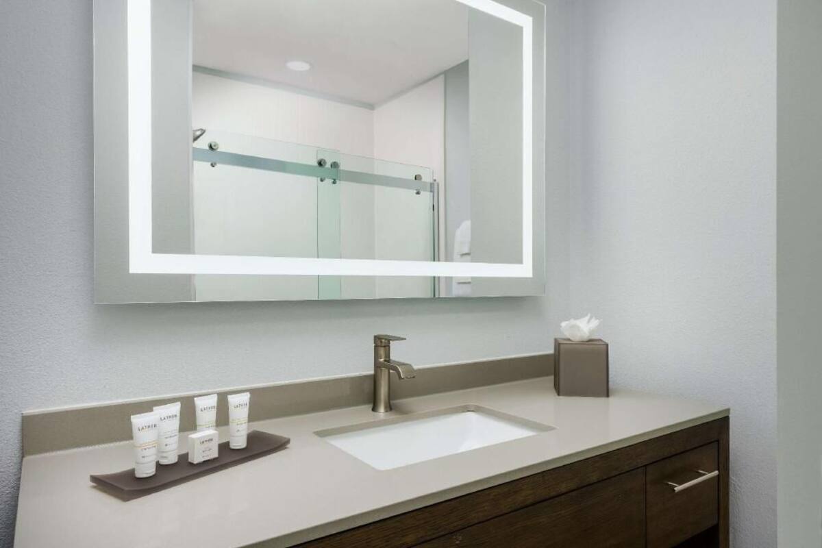 Un baño con un espejo grande, un lavabo, un mueble de tocador con cajones y un estante con artículos de aseo.