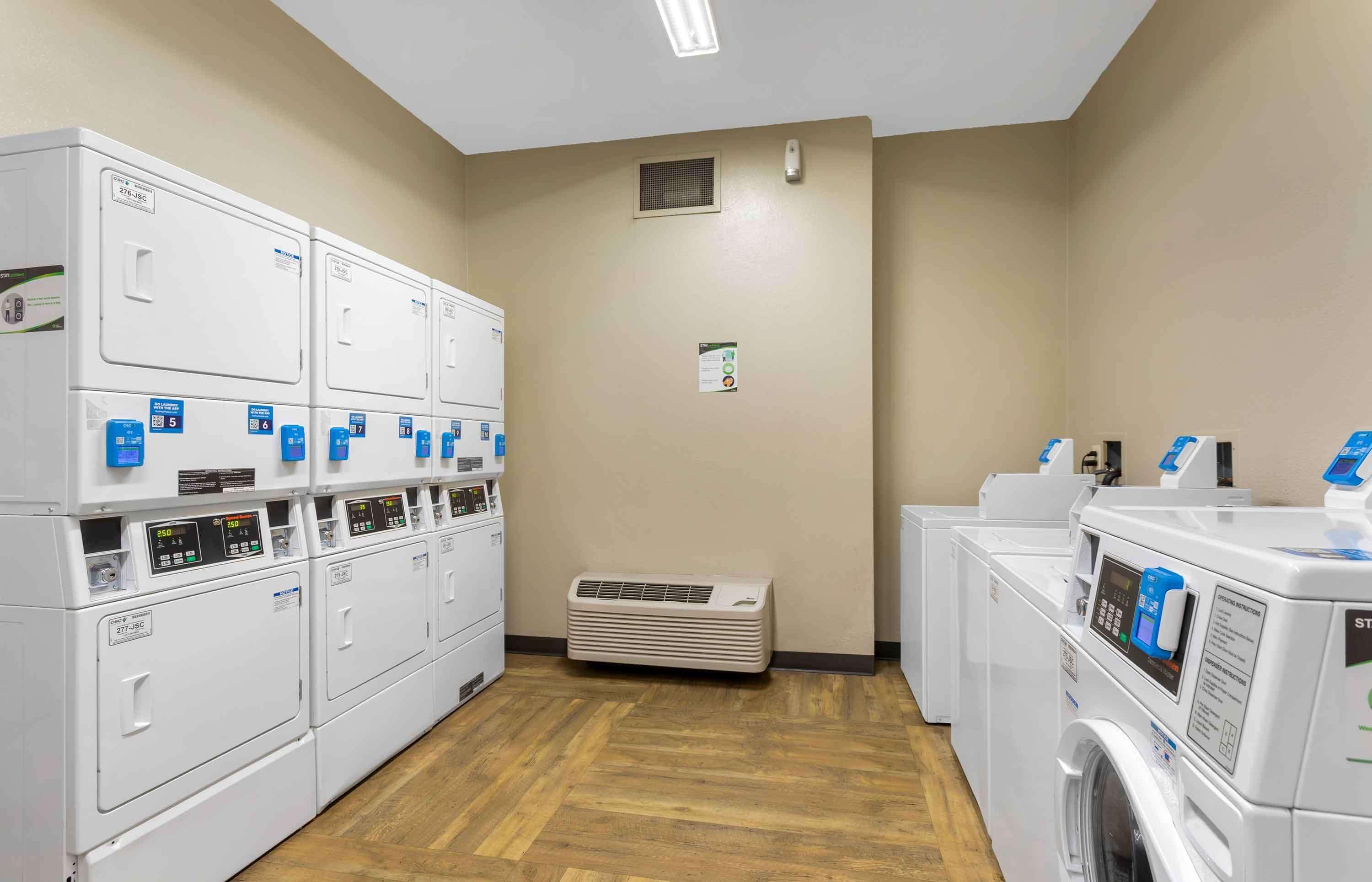 A laundry facility with multiple washing machines and dryers, a vent, and a wall-mounted air conditioning unit.