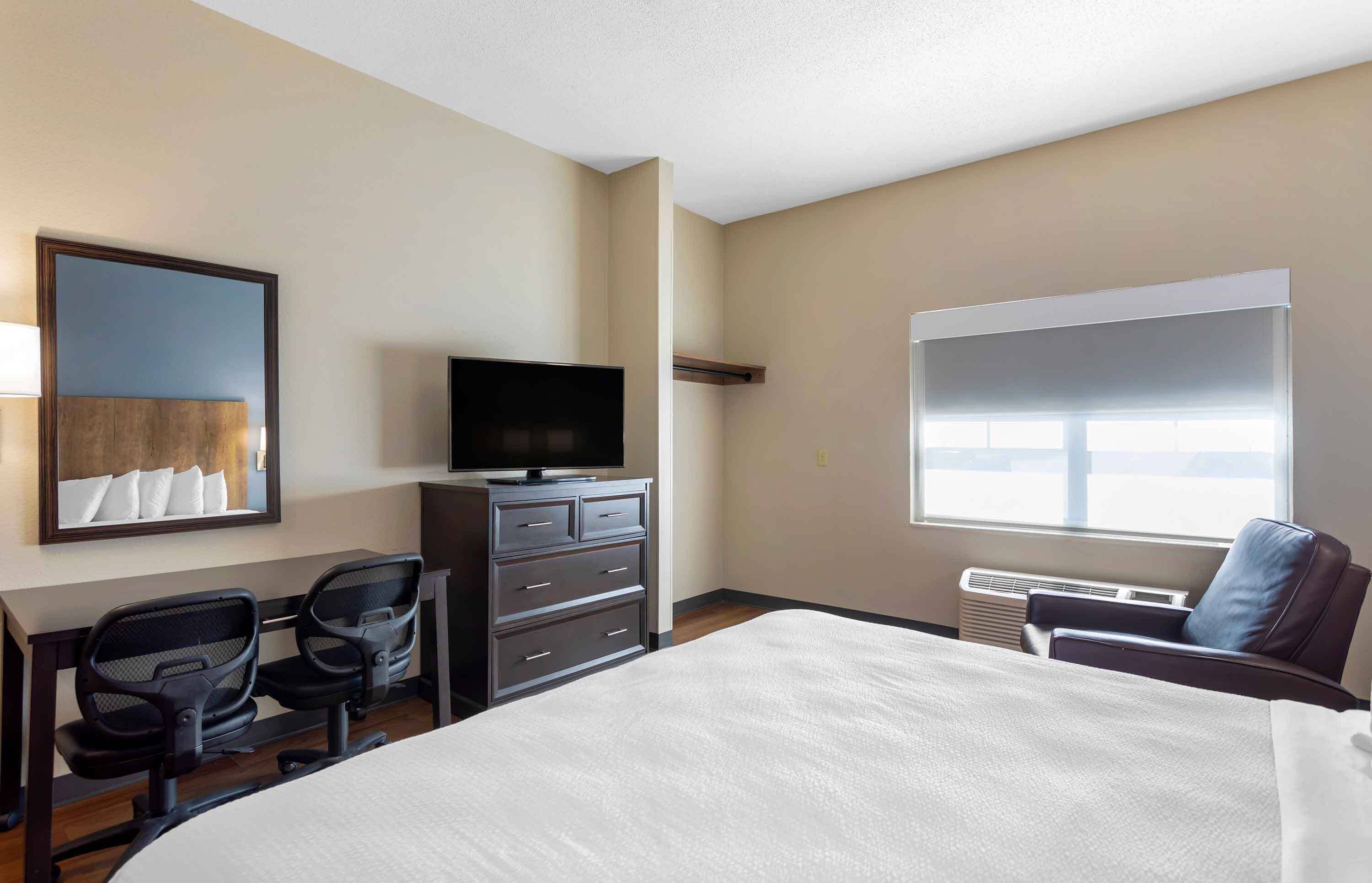 A hotel room with a bed, a TV, a desk with a chair, a window, and a wall-mounted mirror.