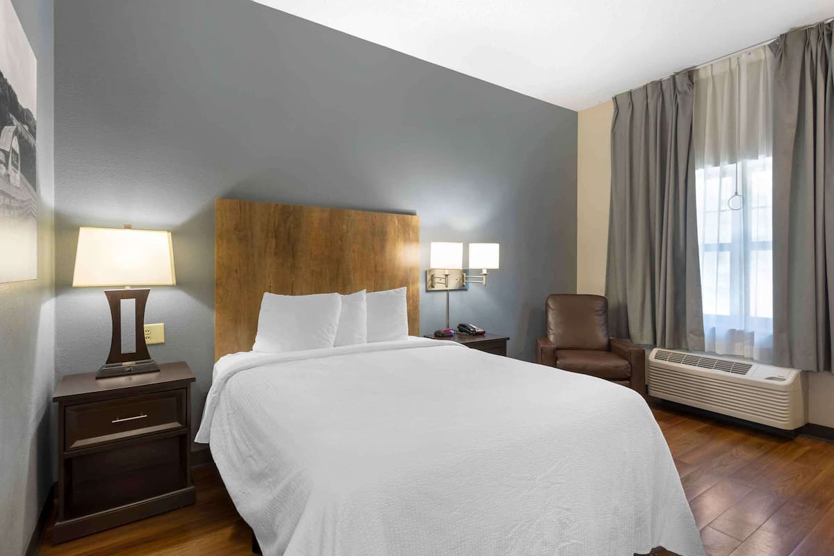 Une chambre d’hôtel avec un grand lit, une tête de lit en bois, deux tables de chevet avec des lampes, une chaise et un appareil de climatisation.