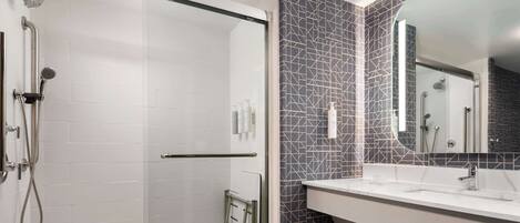 Un baño moderno con ducha envuelta en vidrio, un lavamanos blanco y un espejo con un salpicadero de azulejos geométricos.