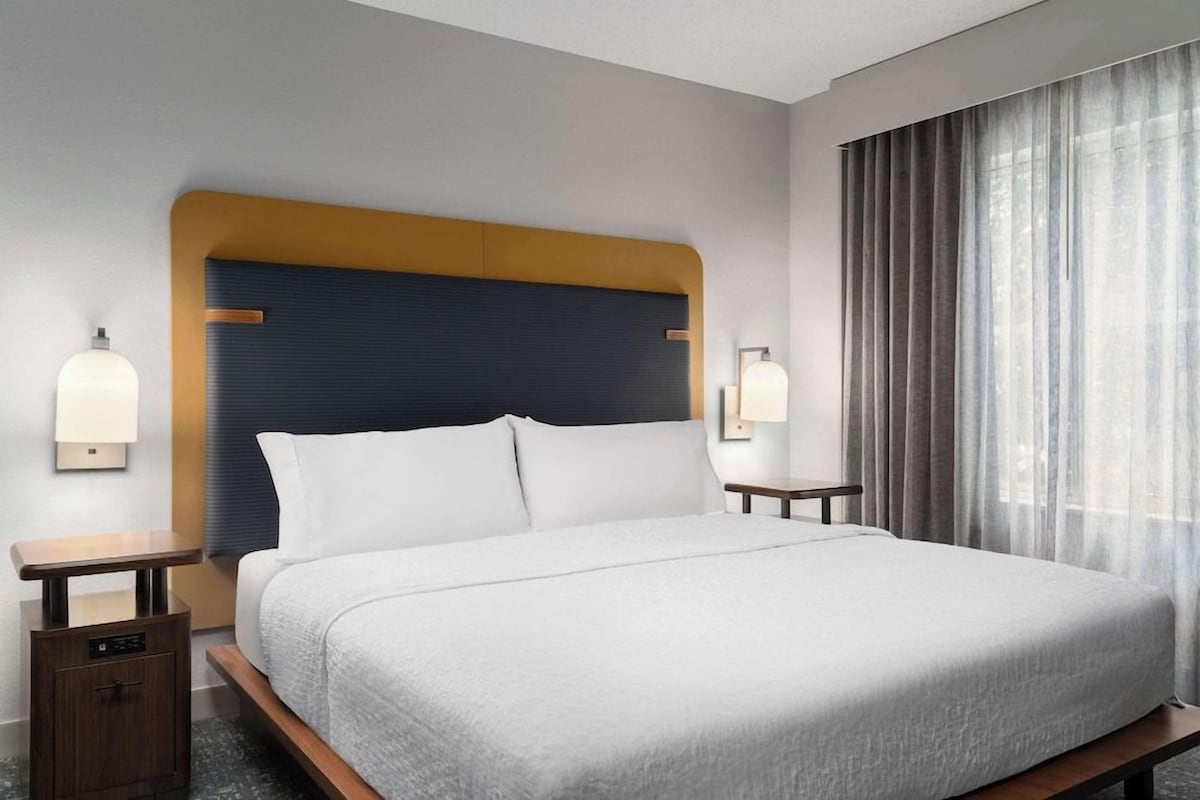 Una habitación de hotel con cama, mesitas de noche y un ventanal con cortinas.