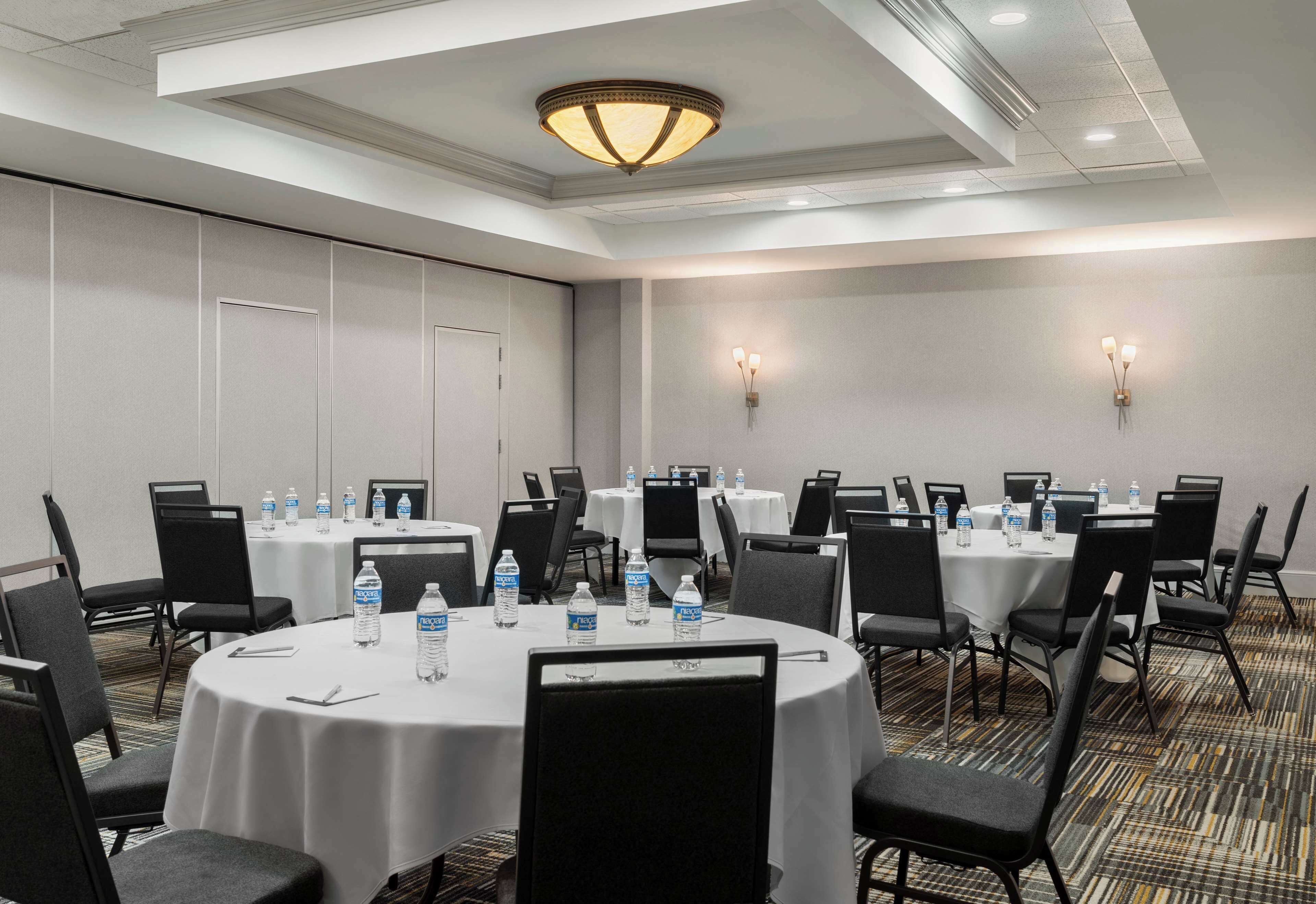 A conference room with round tables, black chairs, white tablecloths, water bottles, and wall-mounted lights.