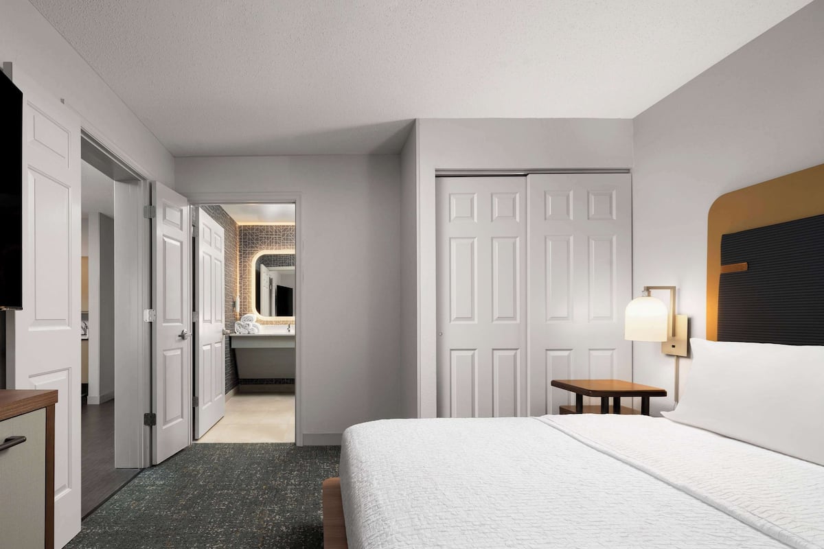 A hotel room with a bed, a desk, a chair, a TV, and a bathroom visible through an open door.