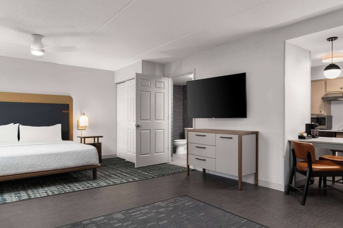 A modern hotel room with a bed, a desk, a chair, a TV, and a bathroom.