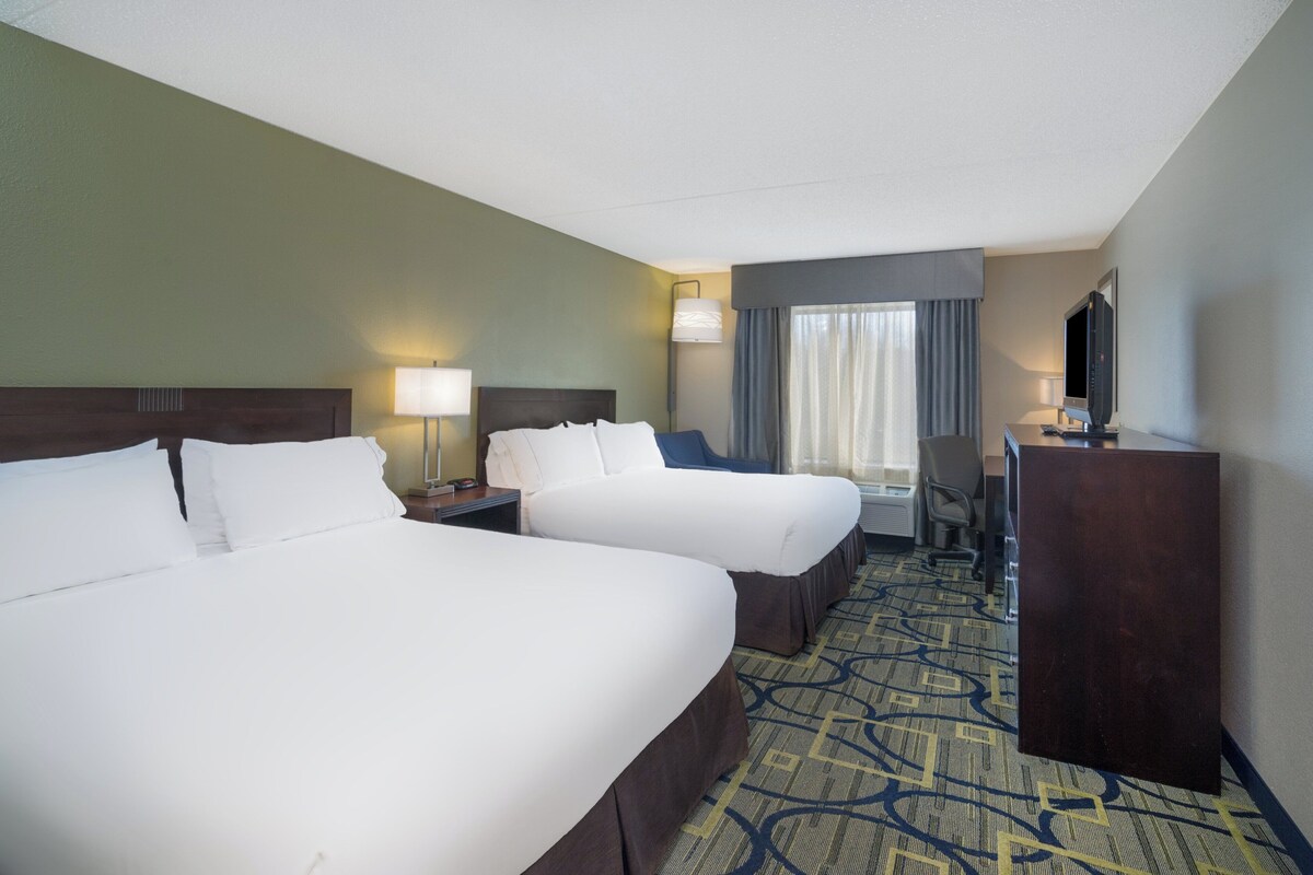 A hotel room with two beds, a desk, a TV, and a window with curtains.