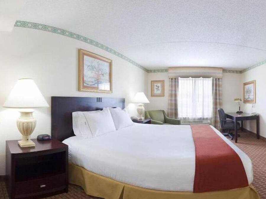A hotel room with a large bed, two bedside tables with lamps, a desk with a chair, a television, and framed artwork on the walls.