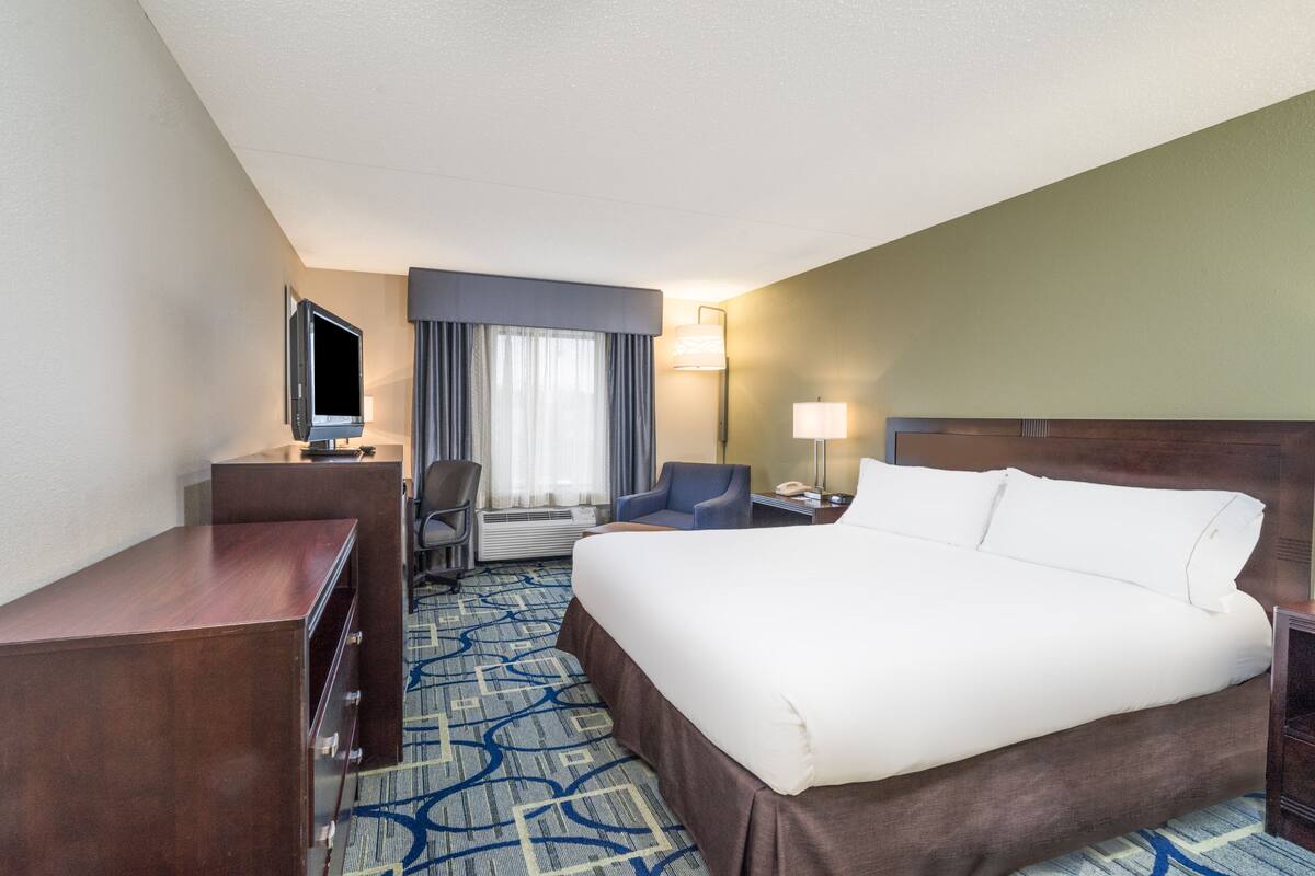 Habitación de hotel con una cama grande, un escritorio con televisor de pantalla plana, una silla, una lámpara de escritorio y una ventana con cortinas.