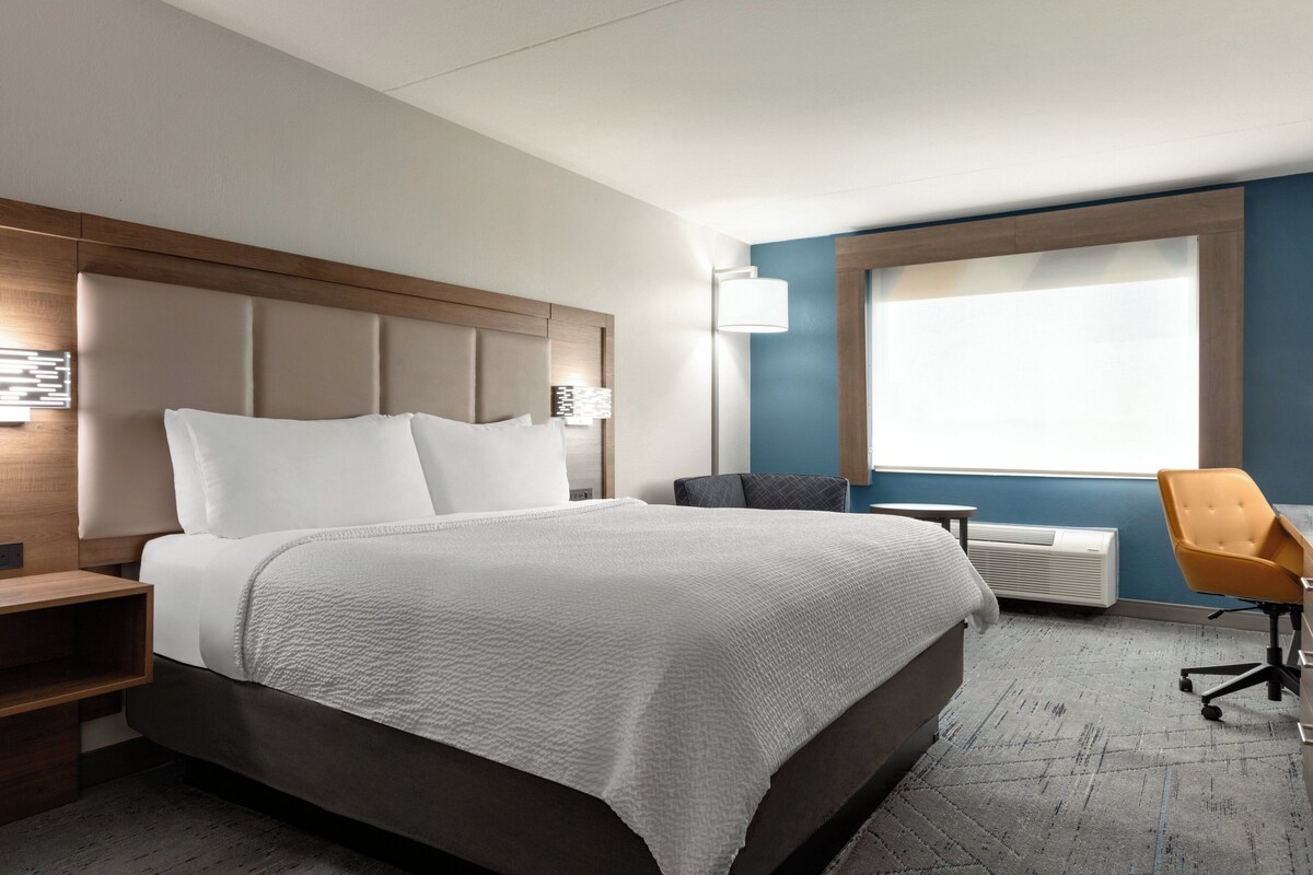 A hotel room with a large bed, a desk, a chair, a window, and a wall-mounted air conditioner.