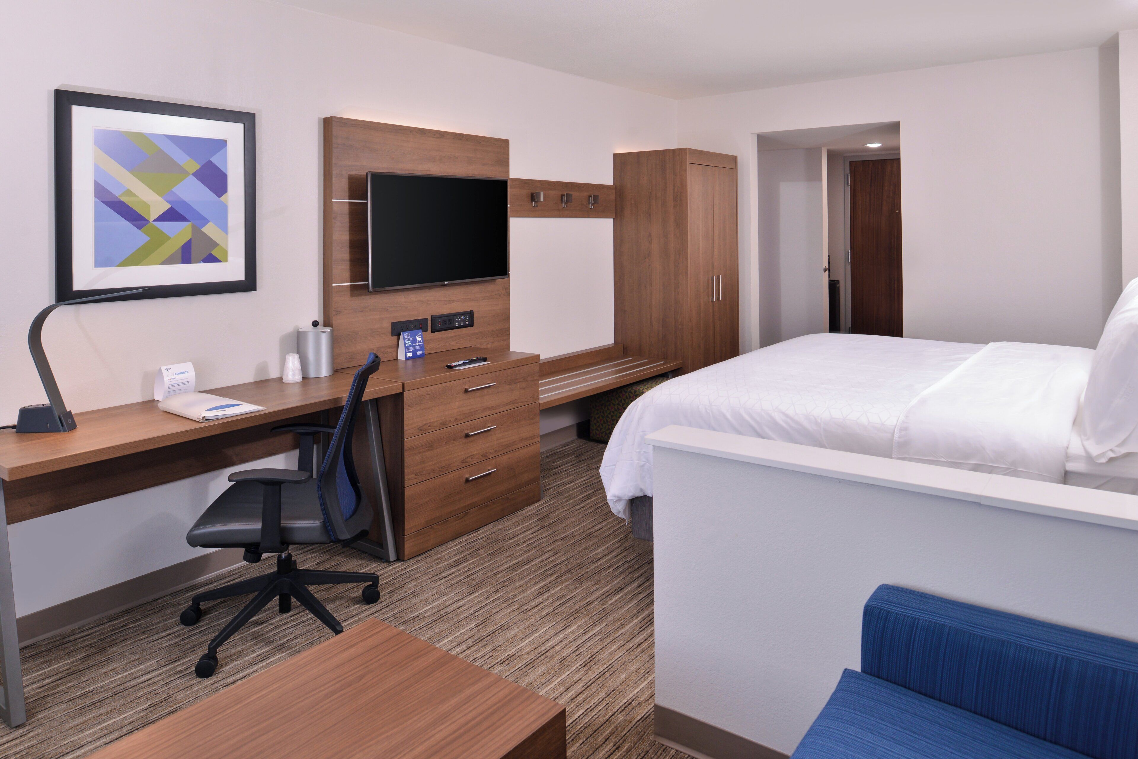 Habitación de hotel con cama, escritorio con silla, televisor, armario y un cuadro en la pared.