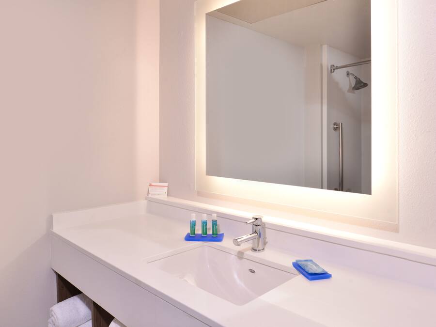 Une salle de bain avec un lavabo blanc, un miroir et une douche que l’on aperçoit dans le reflet.