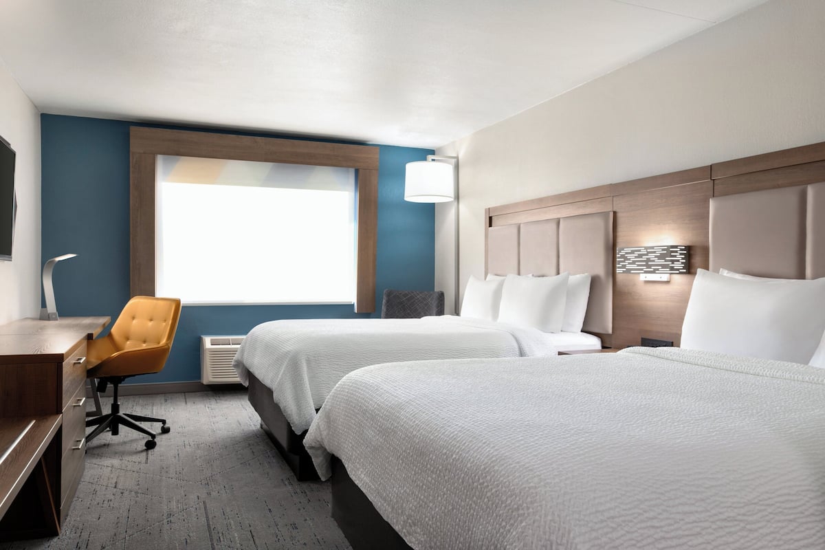 A hotel room with two beds, a desk, a chair, a window, and a wall-mounted lamp.