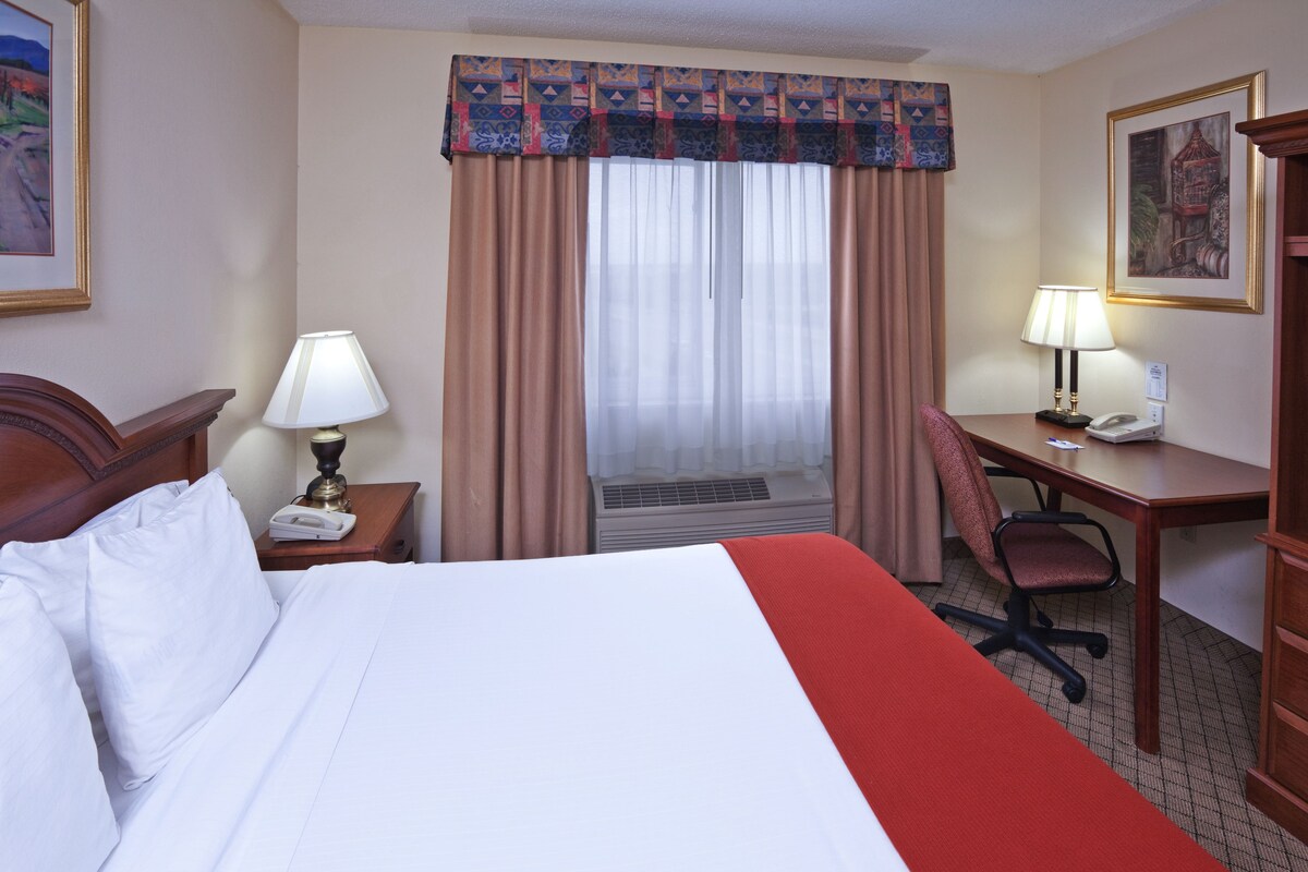 A hotel room with a bed, a desk, a chair, a lamp, and a painting on the wall.