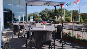 Un espace extérieur aménagé avec des meubles en osier et des plantes en pot, offrant une vue sur un parking et un bâtiment à la façade en verre.