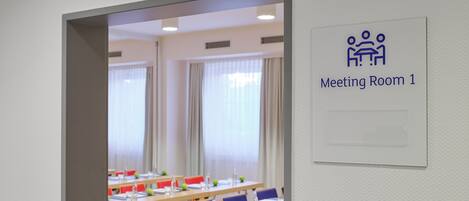 Una sala de reuniones con mesas y sillas, un cartel que indica que es la Sala de Reuniones 1 y una ventana con cortinas.