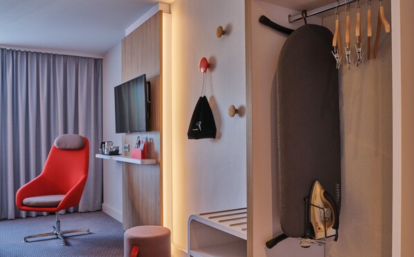 Ein modernes Hotelzimmer mit einem Flachbildfernseher, einem roten Sessel, einem kleinen Tisch mit einem roten Gegenstand und einem Bügelbrett mit Kleiderbügeln.